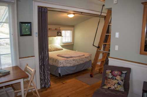 Cozy bedroom with a bed, ladder, and a small dining area, featuring wooden floors and natural light.