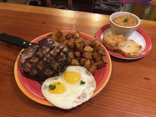 A hearty meal featuring a grilled steak, fried eggs, seasoned potatoes, and a bowl of soup with biscuits on the side.