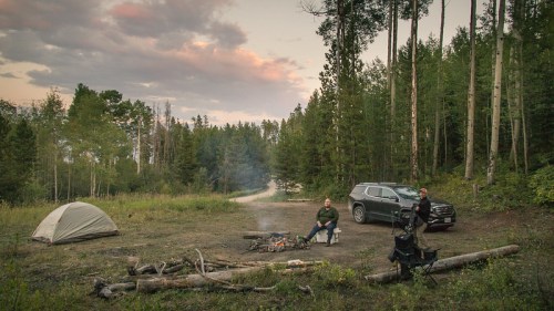 Two campers sit by a fire near a tent and SUV, surrounded by trees and a scenic sunset.