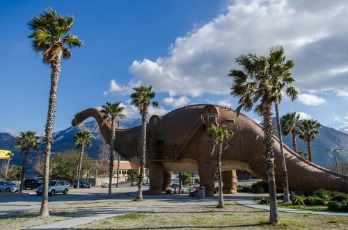 A large dinosaur sculpture surrounded by palm trees, set against a backdrop of mountains and blue sky.