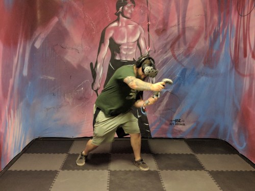 A person wearing a VR headset and controllers stands in a colorful room with a mural of a muscular figure on the wall.