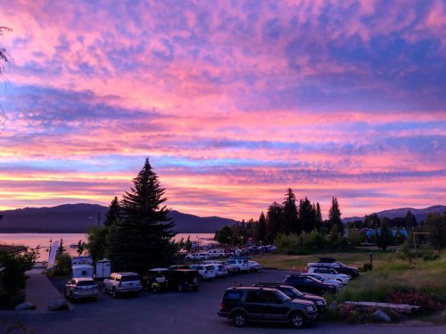 A vibrant sunset paints the sky in pink and purple hues over a lake, with parked cars and trees in the foreground.