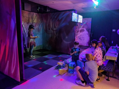 A child in a VR headset plays a game while others watch and sit nearby, surrounded by colorful lights and decorations.