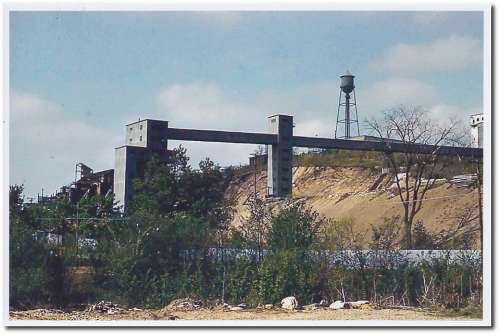 A concrete structure on a hillside with a water tower in the background, surrounded by trees and shrubs.