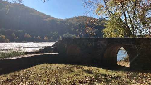 A stone arch bridge near a river, surrounded by trees and hills, with autumn foliage and sunlight filtering through.