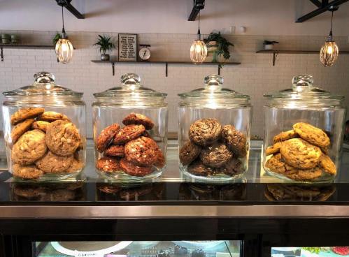Four glass jars filled with assorted cookies, including chocolate chip and oatmeal, displayed on a counter.