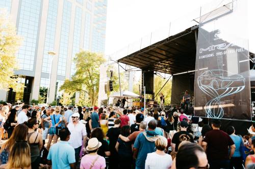 A large crowd enjoys a live music performance at an outdoor festival, with a stage and city buildings in the background.