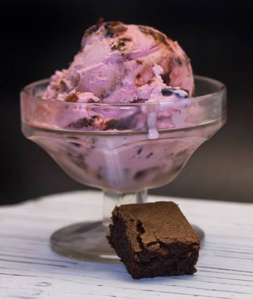 A glass dish filled with pink ice cream topped with chocolate chunks, accompanied by a small piece of brownie.