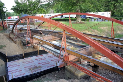 A red metal arch bridge under construction, with supports and a gravel base, surrounded by trees and trucks in the background.