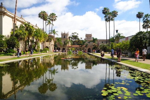 A serene park scene with a reflecting pond, palm trees, and people strolling near historic buildings under a blue sky.