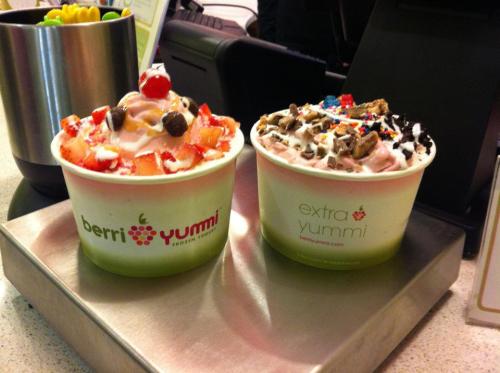 Two colorful frozen yogurt cups topped with strawberries, chocolate chips, sprinkles, and cherries, sitting on a counter.