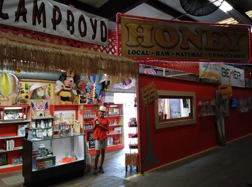 A vendor booth at a market featuring honey products, colorful signage, and a person in an orange shirt.