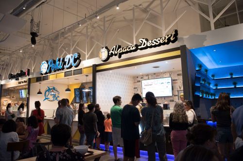 A bustling food hall with people lined up at various food stalls, including "Poki DC" and "Alpaca Dessert."