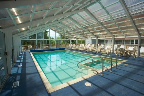 Indoor swimming pool with a glass roof, surrounded by lounge chairs and a blue pool cover. Bright and spacious atmosphere.