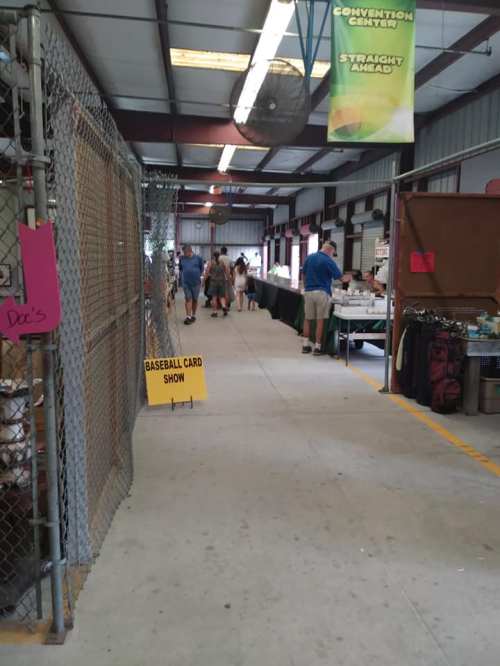 Interior of a market with vendors, a sign for a baseball card show, and people browsing tables.