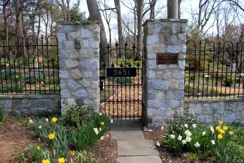 A stone gate with a black iron fence, leading into a garden with blooming flowers and a sign reading "The Wishing Cemetery."