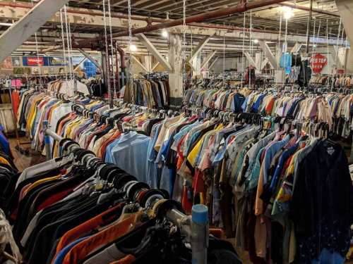 A large thrift store with rows of colorful shirts and clothing hanging on racks under bright lights.