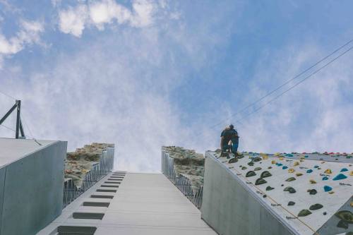 A climber ascends a tall rock wall under a blue sky, with a second wall visible on the left.