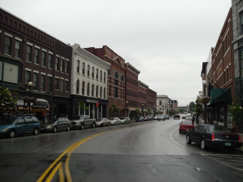 A rainy street scene featuring historic buildings, parked cars, and trees lining the sidewalk.