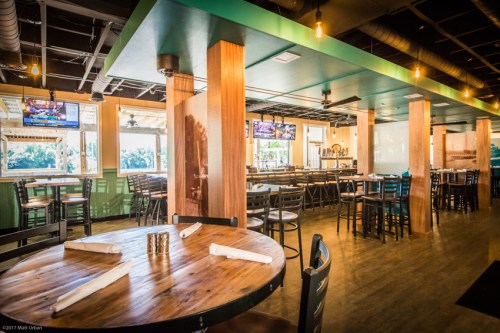 Interior of a modern restaurant featuring wooden tables, bar seating, and multiple TVs on the walls.