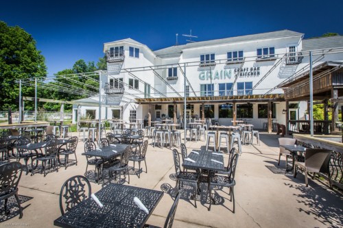 Outdoor seating area of a restaurant with black tables and chairs, surrounded by greenery and a white building in the background.