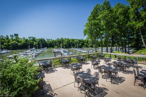 Outdoor dining area with black tables overlooking a marina filled with boats, surrounded by lush green trees.