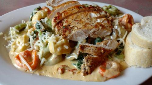 A plate of creamy pasta with grilled chicken, shrimp, and vegetables, served with a slice of bread.