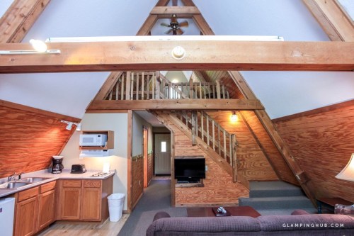 Cozy A-frame interior with wooden beams, kitchen area, living space, and staircase leading to upper level.