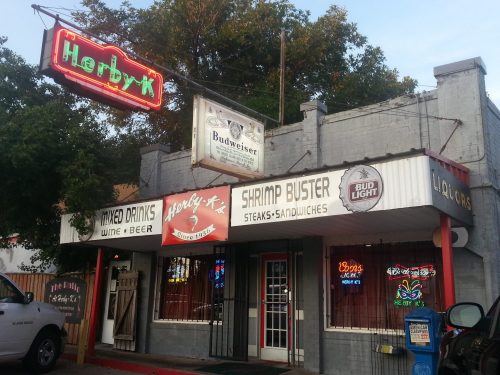 A colorful restaurant exterior featuring signs for drinks, shrimp, and sandwiches, with neon lights and a welcoming entrance.