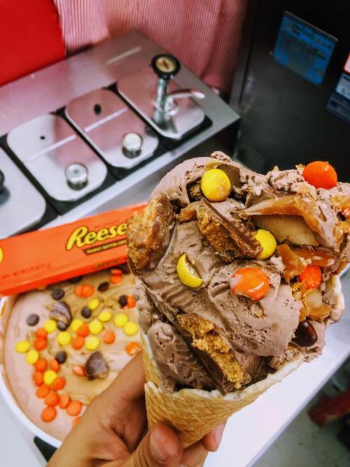 A hand holds a cone filled with chocolate ice cream topped with Reese's pieces and chunks, with more ice cream in the background.
