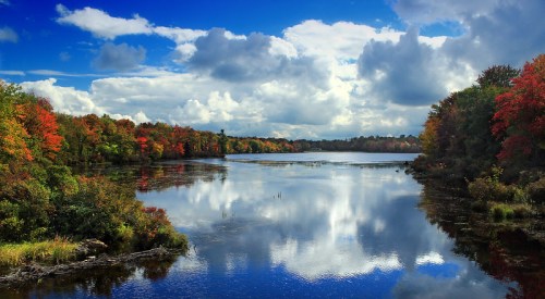 A serene lake surrounded by vibrant autumn foliage and a blue sky with fluffy clouds reflecting on the water.