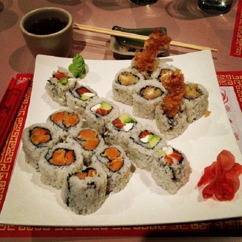 A plate of assorted sushi rolls, including tempura, with pickled ginger and a cup of tea in the background.