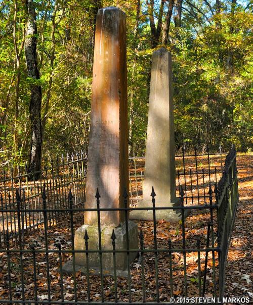 Two weathered gravestones surrounded by a black iron fence, set in a wooded area with fallen leaves.