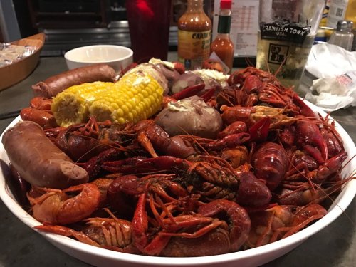 A large bowl filled with crawfish, sausage, corn, and potatoes, accompanied by hot sauce bottles in the background.