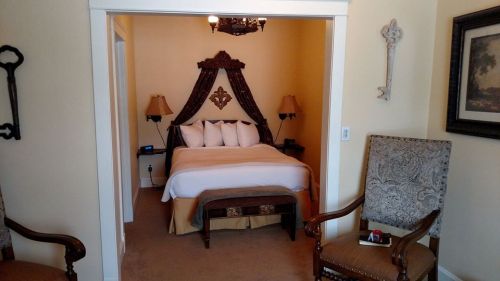 Cozy bedroom with a large bed, decorative headboard, two lamps, and elegant chairs in a warm-toned room.