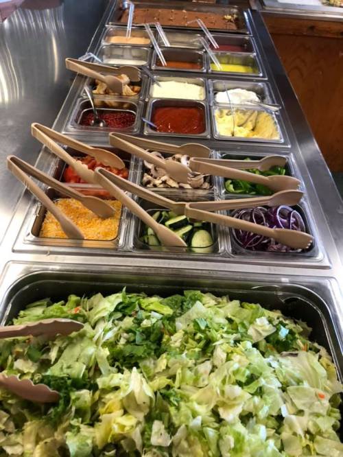 A salad bar with various toppings, including vegetables, dressings, and sauces, arranged in metal containers.
