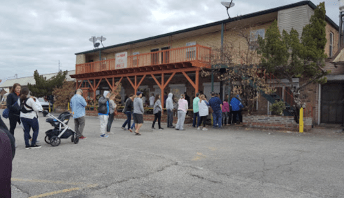 A line of people waiting outside a building with a wooden deck, surrounded by trees and cloudy skies.