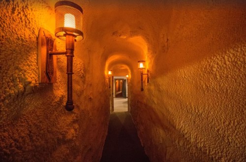 A dimly lit, narrow corridor with textured walls and wall-mounted lanterns, leading to an open doorway.