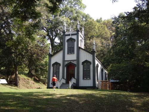 A person walks towards a tall, white church with black trim and spires, surrounded by trees in a grassy area.