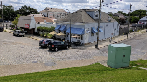 A quaint building with blue awnings, outdoor seating, and parked cars on a cobblestone street under a cloudy sky.
