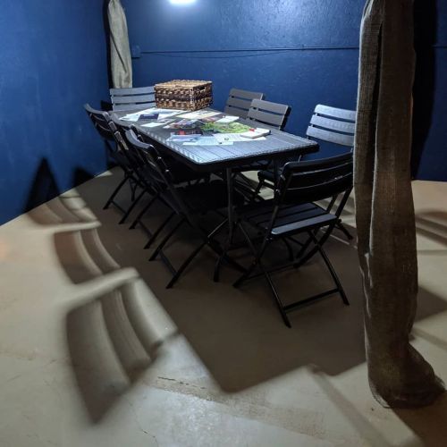 A dimly lit room with a table surrounded by folding chairs and a basket, casting shadows on the floor.