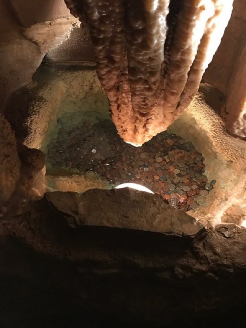 A cave with stalactites above a pool of water filled with colorful coins at the bottom.