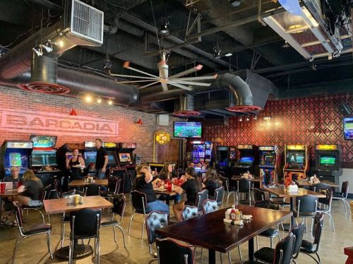 A lively bar with arcade games, patrons dining, and a retro atmosphere featuring exposed brick and industrial decor.