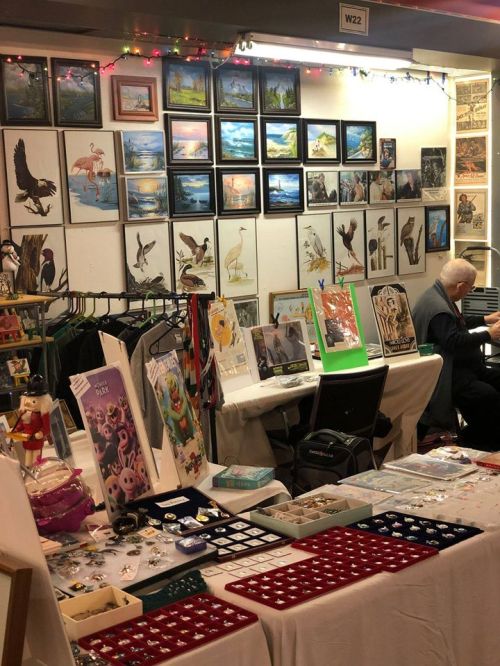 A display of artwork and crafts at a market, with paintings on the wall and various items on tables.