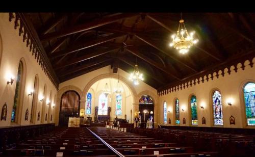 Interior of a church featuring stained glass windows, wooden beams, and rows of pews. Soft lighting enhances the atmosphere.