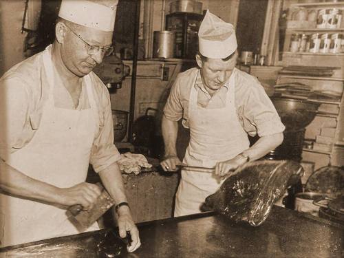 Two men in white aprons and hats work in a kitchen, preparing food with large utensils on a metal surface.