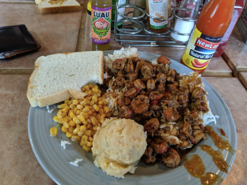 A plate of rice topped with seasoned shrimp, corn, mashed potatoes, two slices of bread, and hot sauce.