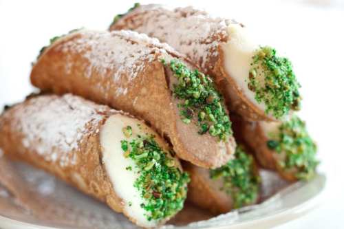 A plate of cannoli filled with creamy filling and topped with green pistachio crumbs and powdered sugar.