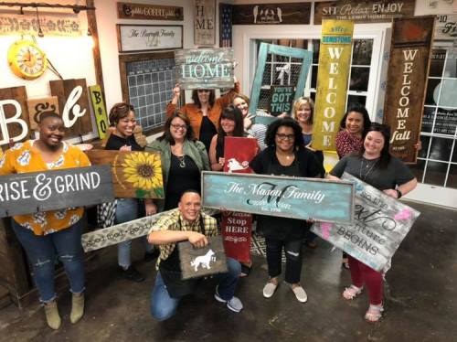 A group of people holding decorative signs in a cozy workshop setting, smiling and posing together.