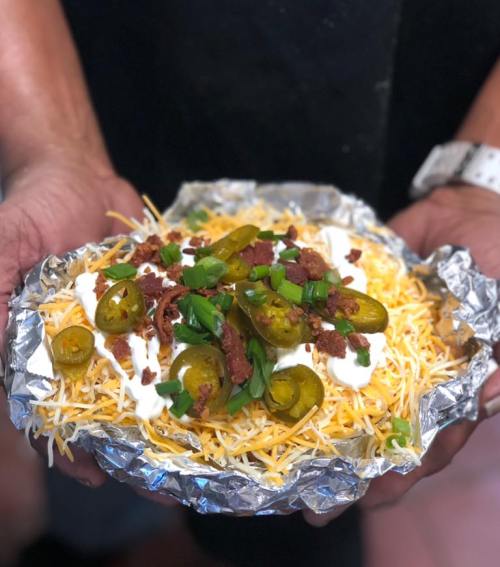 A person holds a foil-wrapped dish topped with cheese, jalapeños, bacon bits, and sour cream.